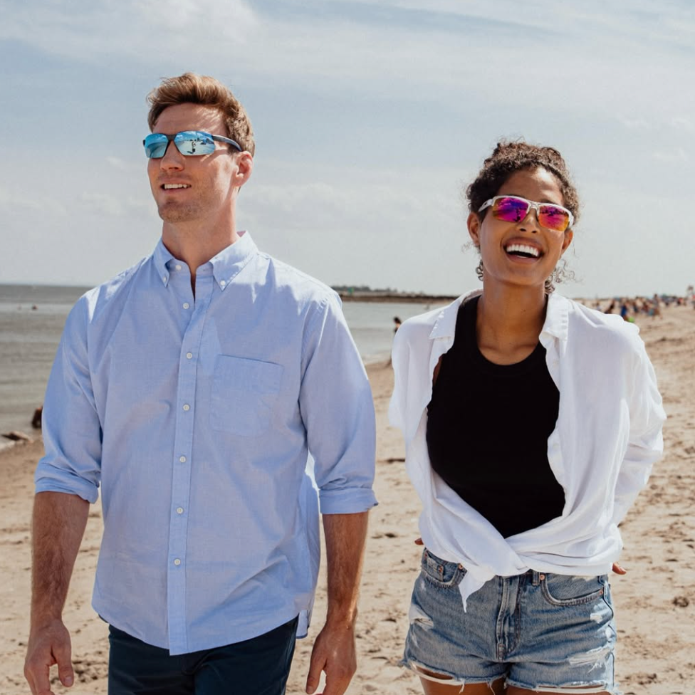 A man and a woman smiling at the beach in sunglasses - with Blue Water lenses and Spectra lenses